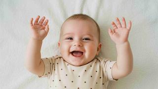 Smiling baby lying on a soft white blanket, arms raised, in a beige polka-dot onesie.
